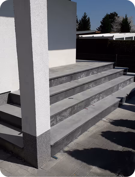 Graue Außentreppe mit vier Stufen neben weißer Wand unter blauem Himmel.