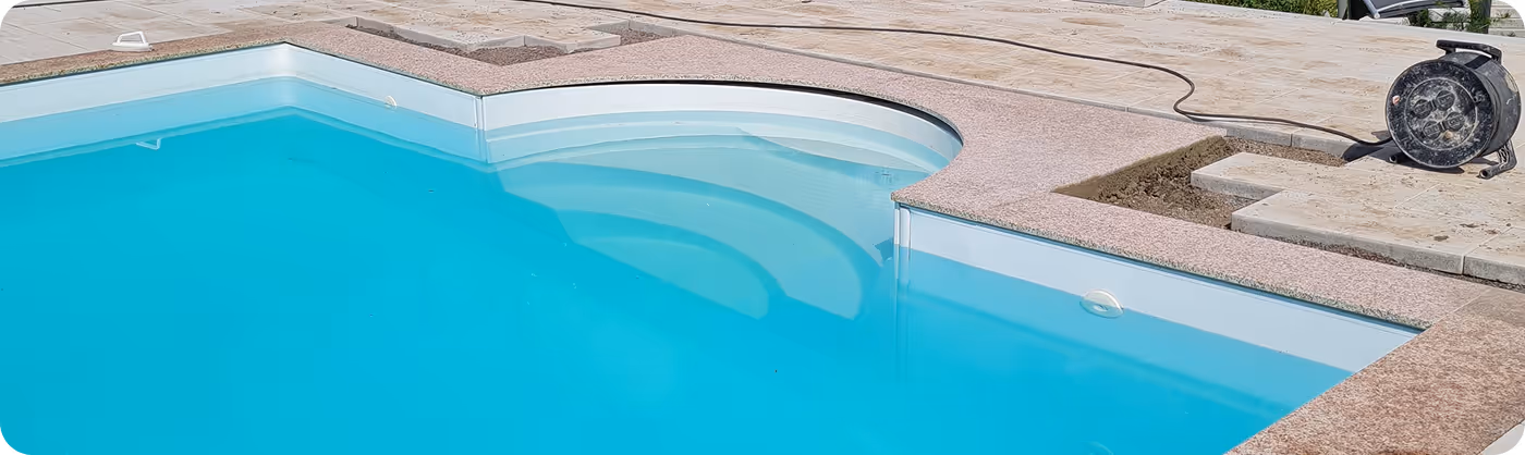Teil eines sauberen blauen Schwimmbeckens mit einer Badeleiter, Marmor- und Steinplatten am Rand, und einer Kabeltrommel auf der Terrasse.