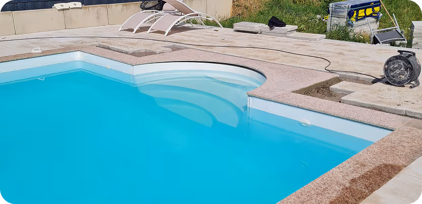 Teil eines blauen Schwimmbeckens mit Treppen und zwei Liegestühlen neben dem Beckenrand auf einer gepflasterten Terrasse.