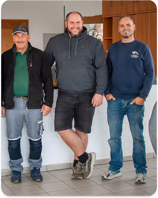 Drei Männer lächeln stehend in einem Innenraum mit heller Wand, der mittlere trägt eine graue Kapuzenjacke und Shorts, die anderen zwei Freizeit- und Arbeitskleidung.